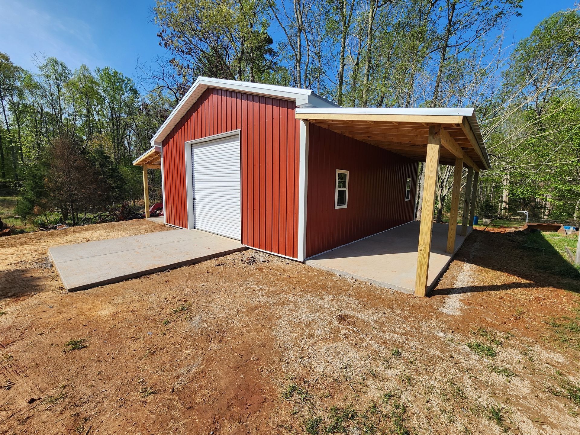 Red metal building with white garage door and covered porch; concrete slab in yard.