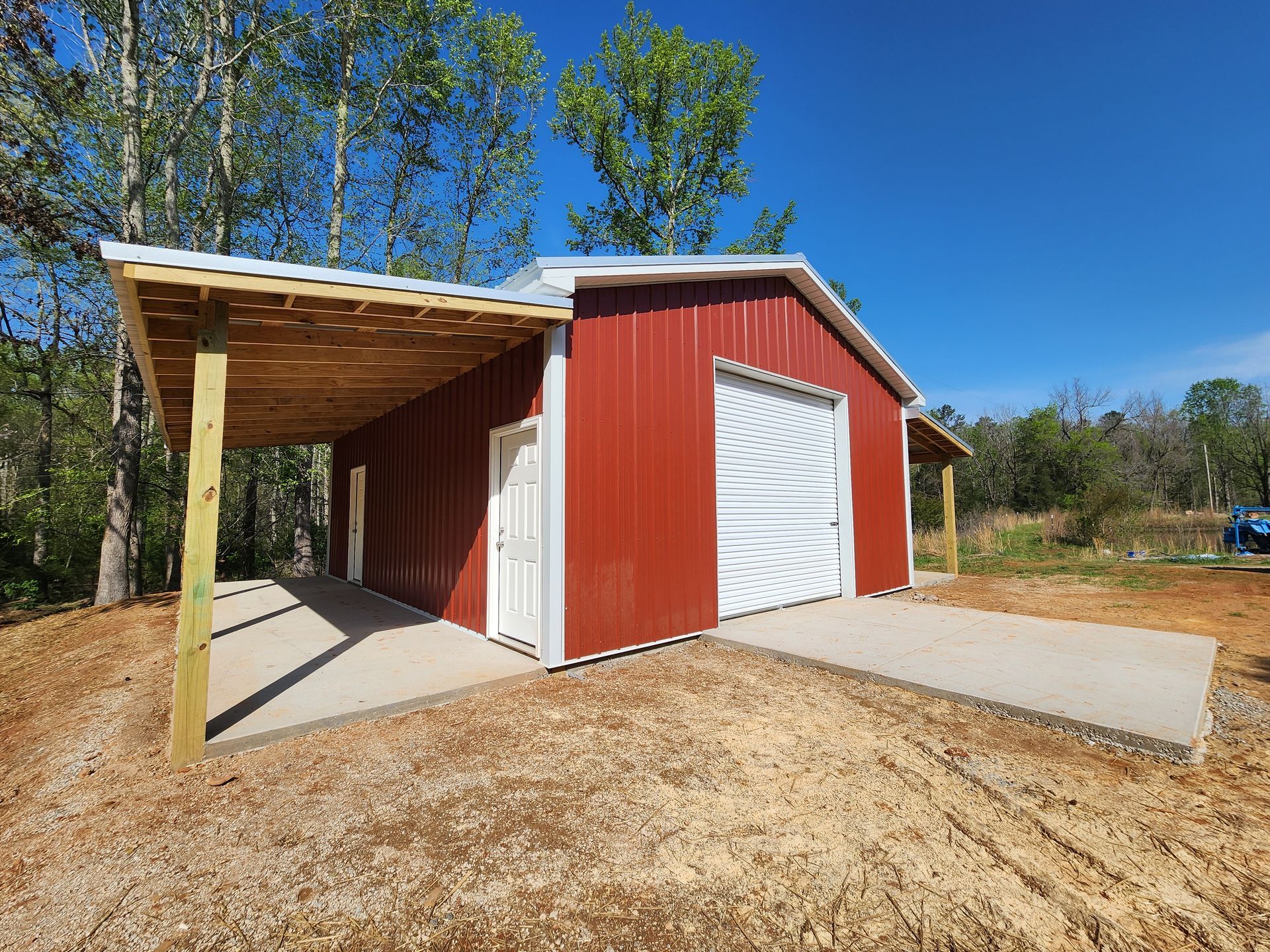 Red metal barn with a concrete slab and carport, surrounded by trees on a sunny day.