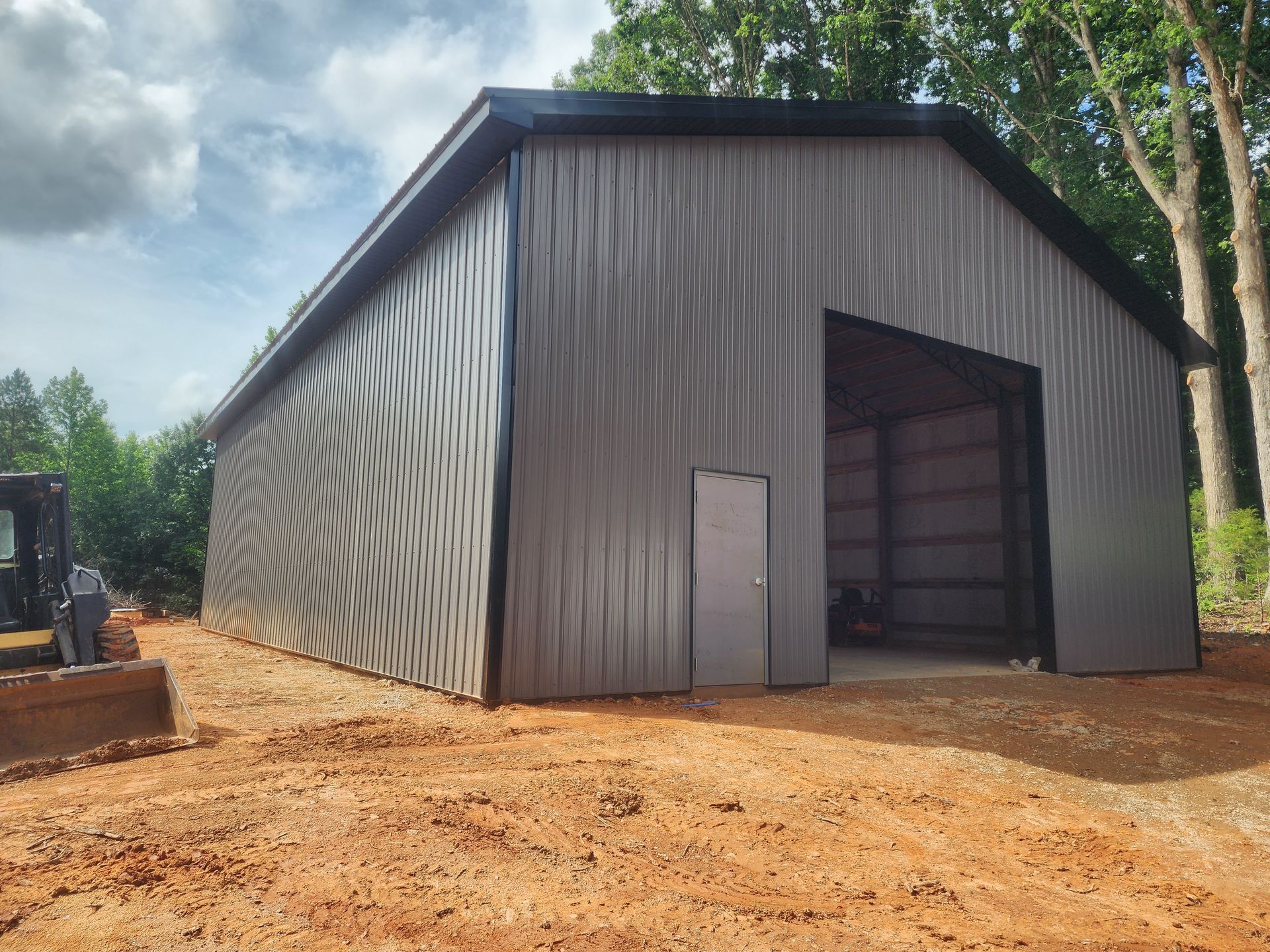 Gray metal barn with a large open doorway, set on a dirt lot, trees in the background.