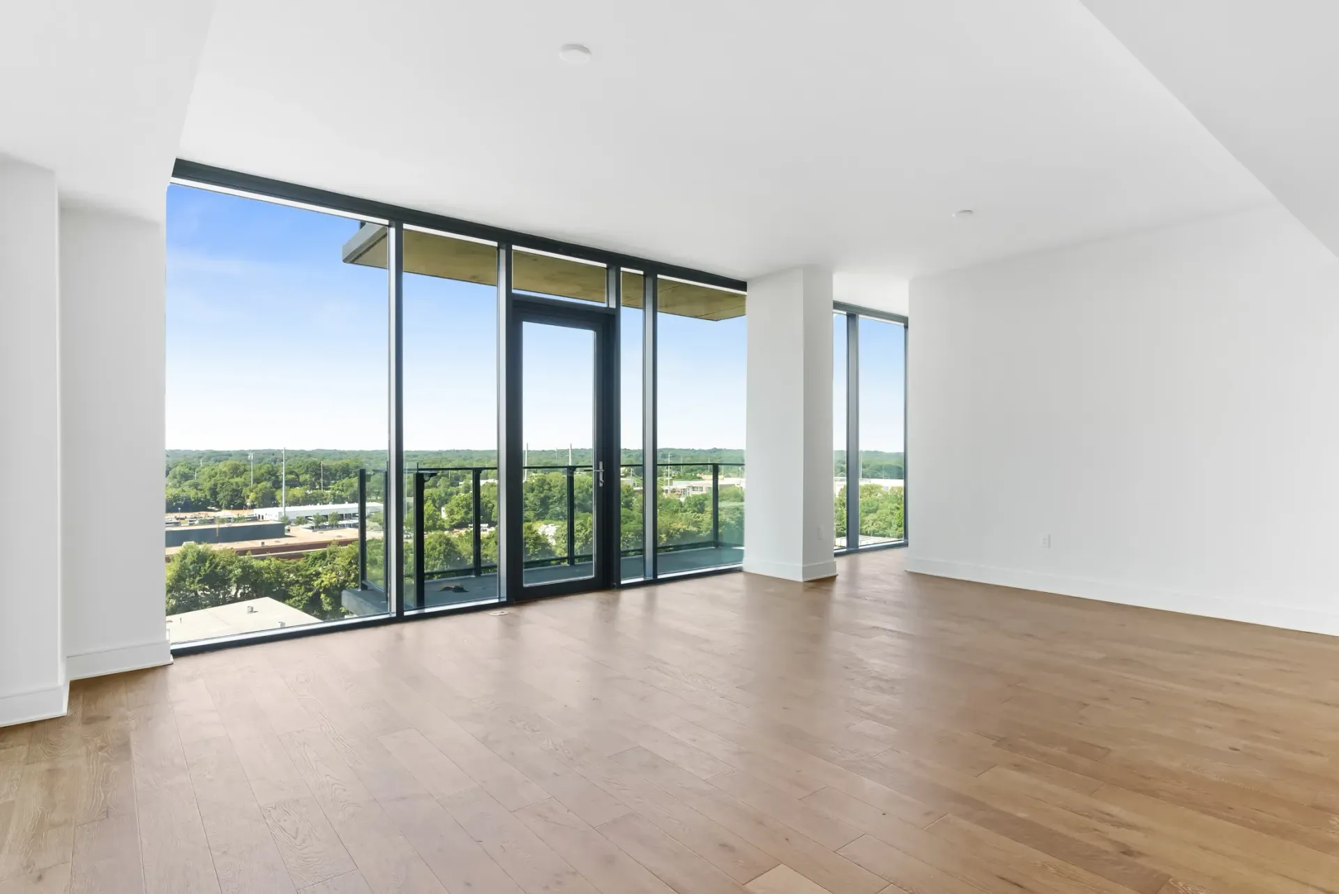 Empty apartment living room with large windows and a balcony overlooking trees and buildings.