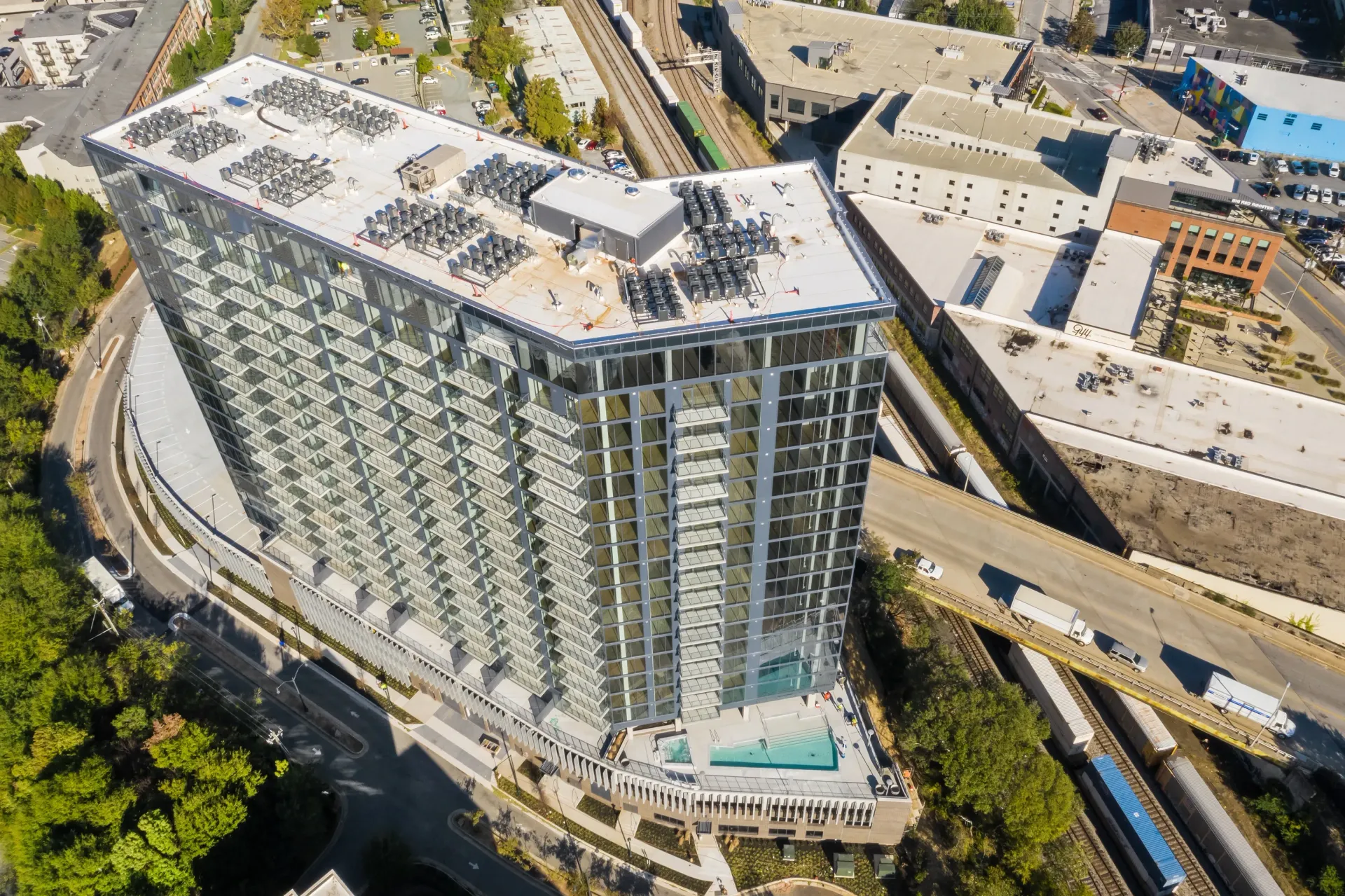 Aerial view of a modern high-rise apartment building with a pool and surrounding city infrastructure.