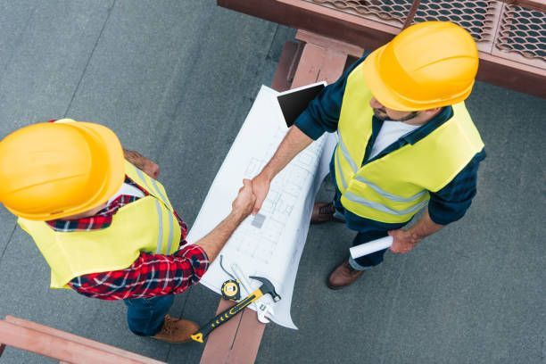 Two construction workers in yellow vests and hard hats shake hands over a blueprint.