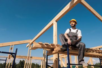 Construction worker in overalls and a hard hat sits on a wooden frame, looking up at the sky.