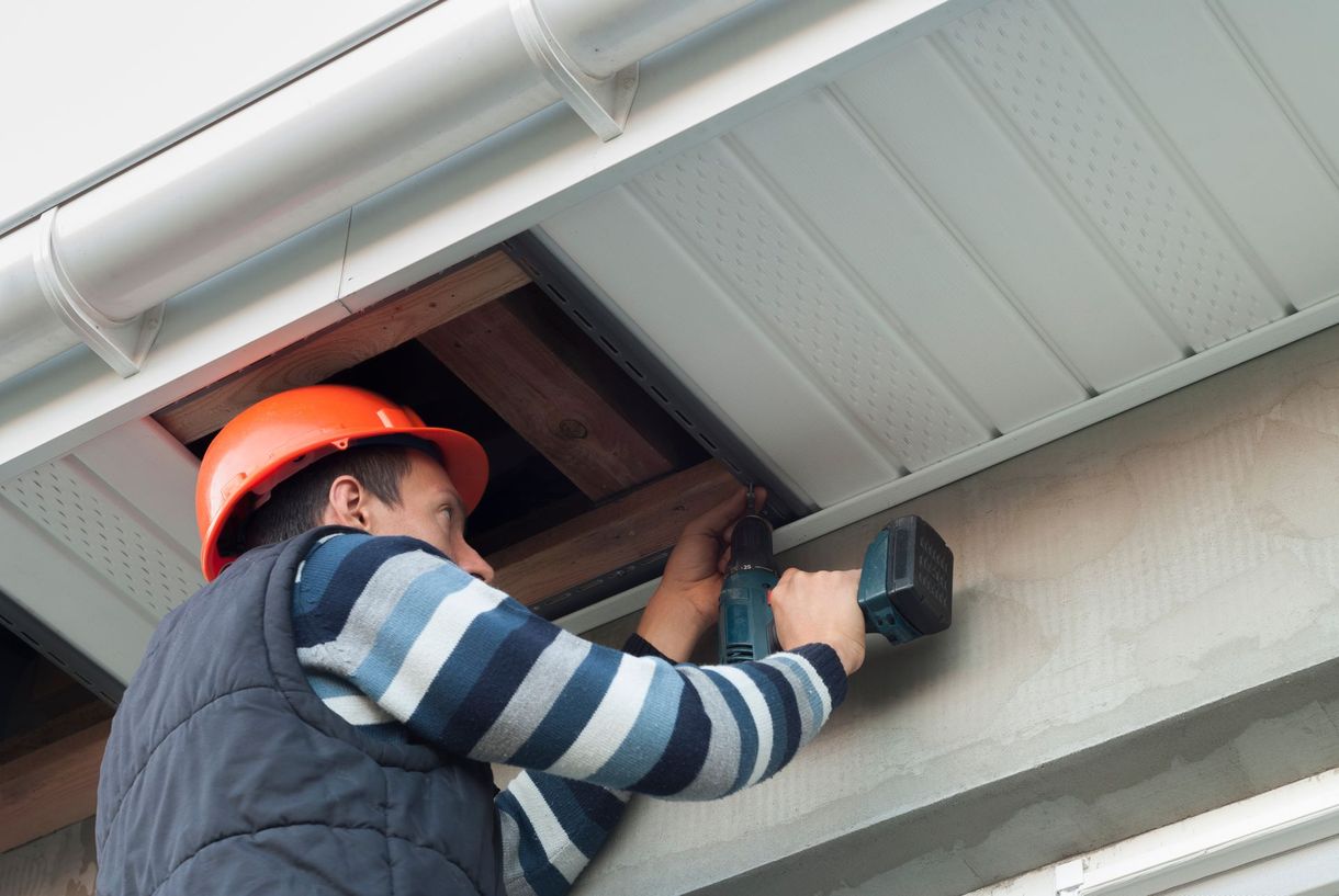 A person in an orange hard hat drills into a white soffit, outdoors near a gutter.