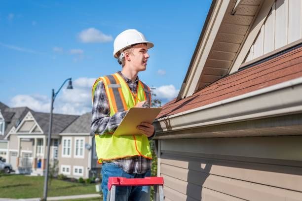 Person in hard hat and vest inspecting a roof with clipboard. Houses and blue sky in background.