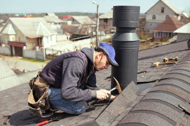 Roofer in blue cap working on a roof, hammering near a black chimney, with houses in the background.