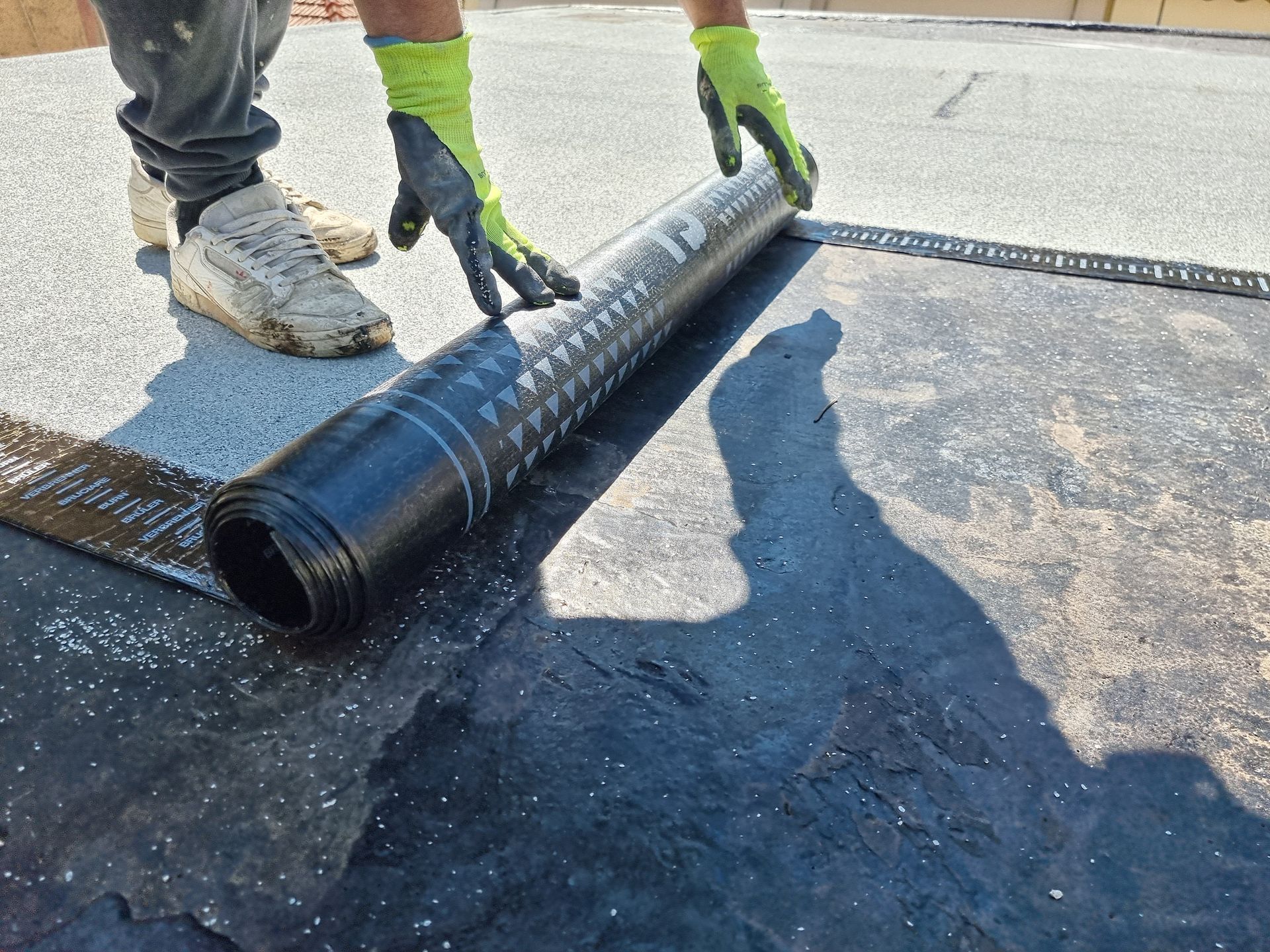 Person rolling out a black roofing membrane on a flat surface, wearing gloves.