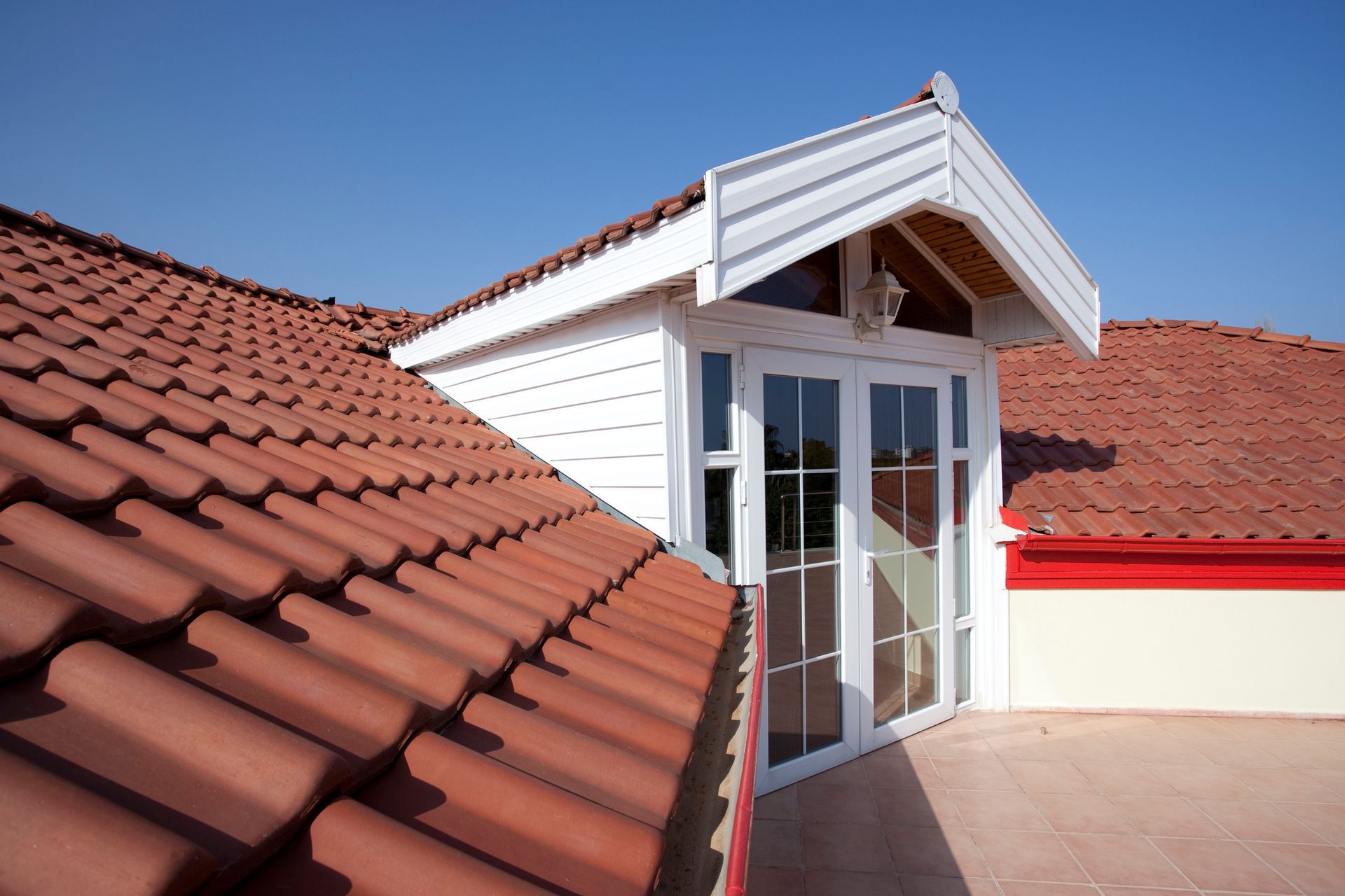 Red-tiled roof with a white dormer featuring double glass doors under a clear blue sky.