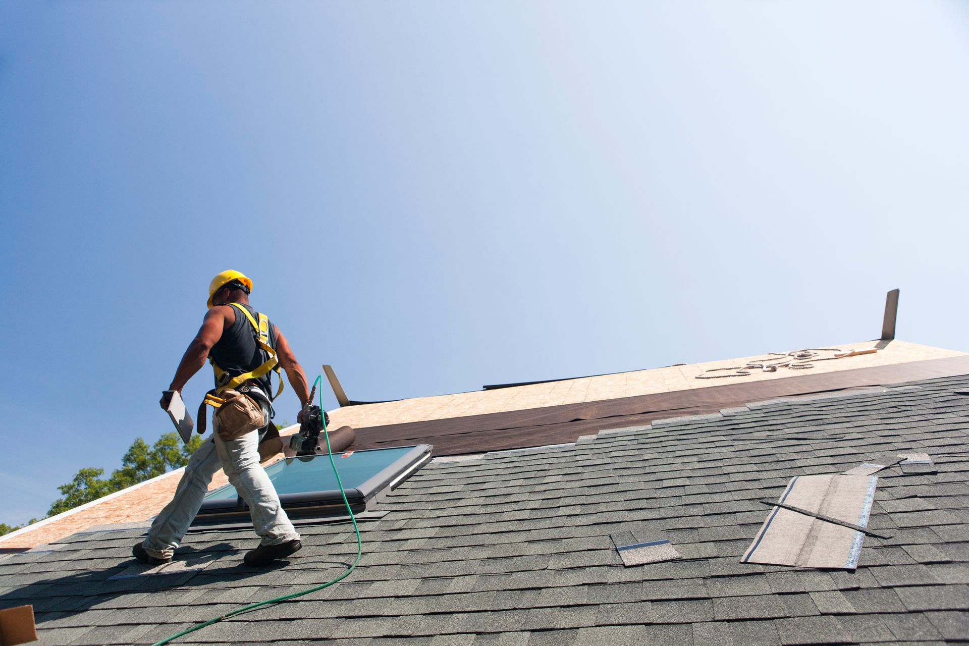 Roofer in safety gear walks on a shingled roof under a clear blue sky.