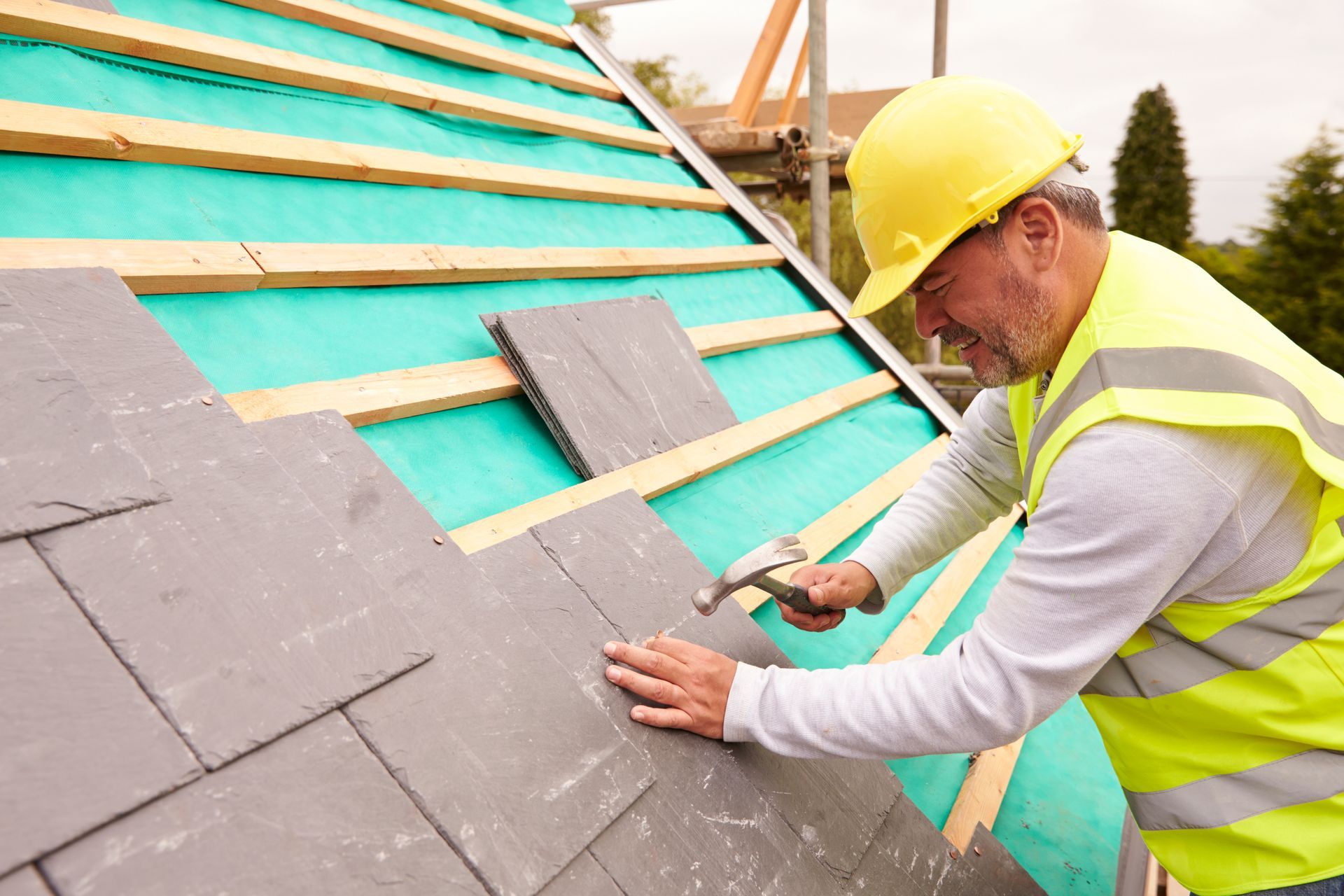 Roofer hammering slate tiles on a roof. Wearing yellow hard hat and vest.