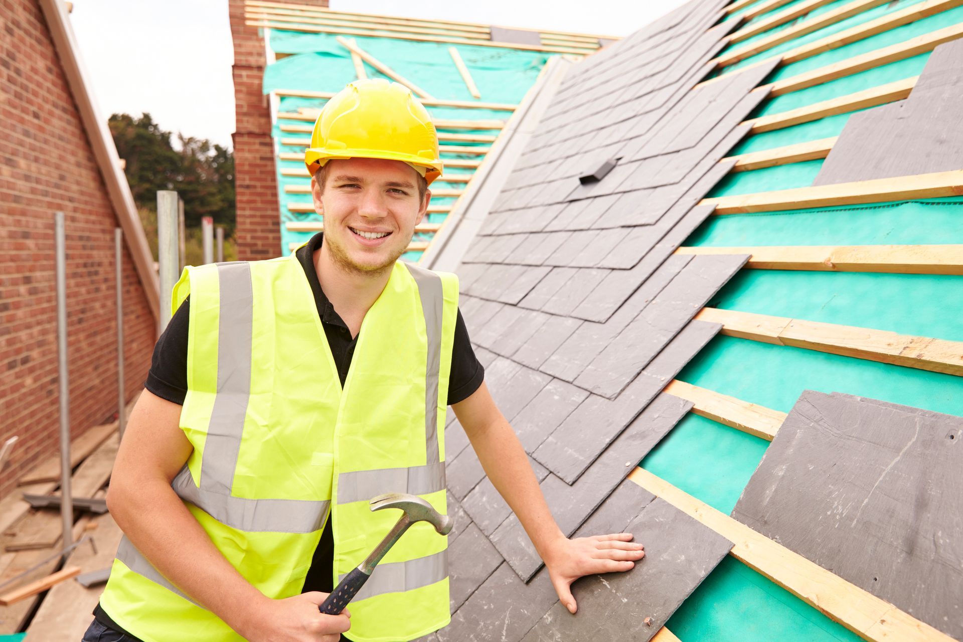 Construction worker on a roof, smiling, installing roofing tiles with hammer. Yellow hard hat and safety vest.