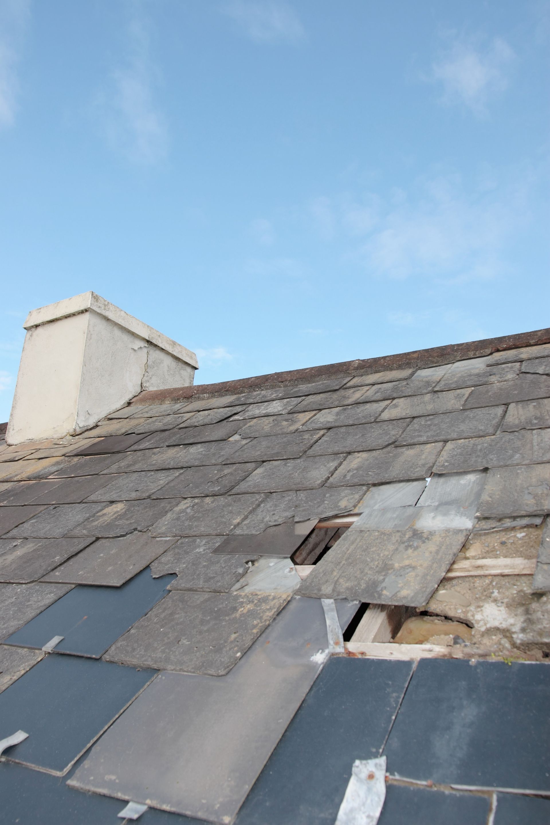 Damaged slate roof with missing tiles, a chimney, and a blue sky.