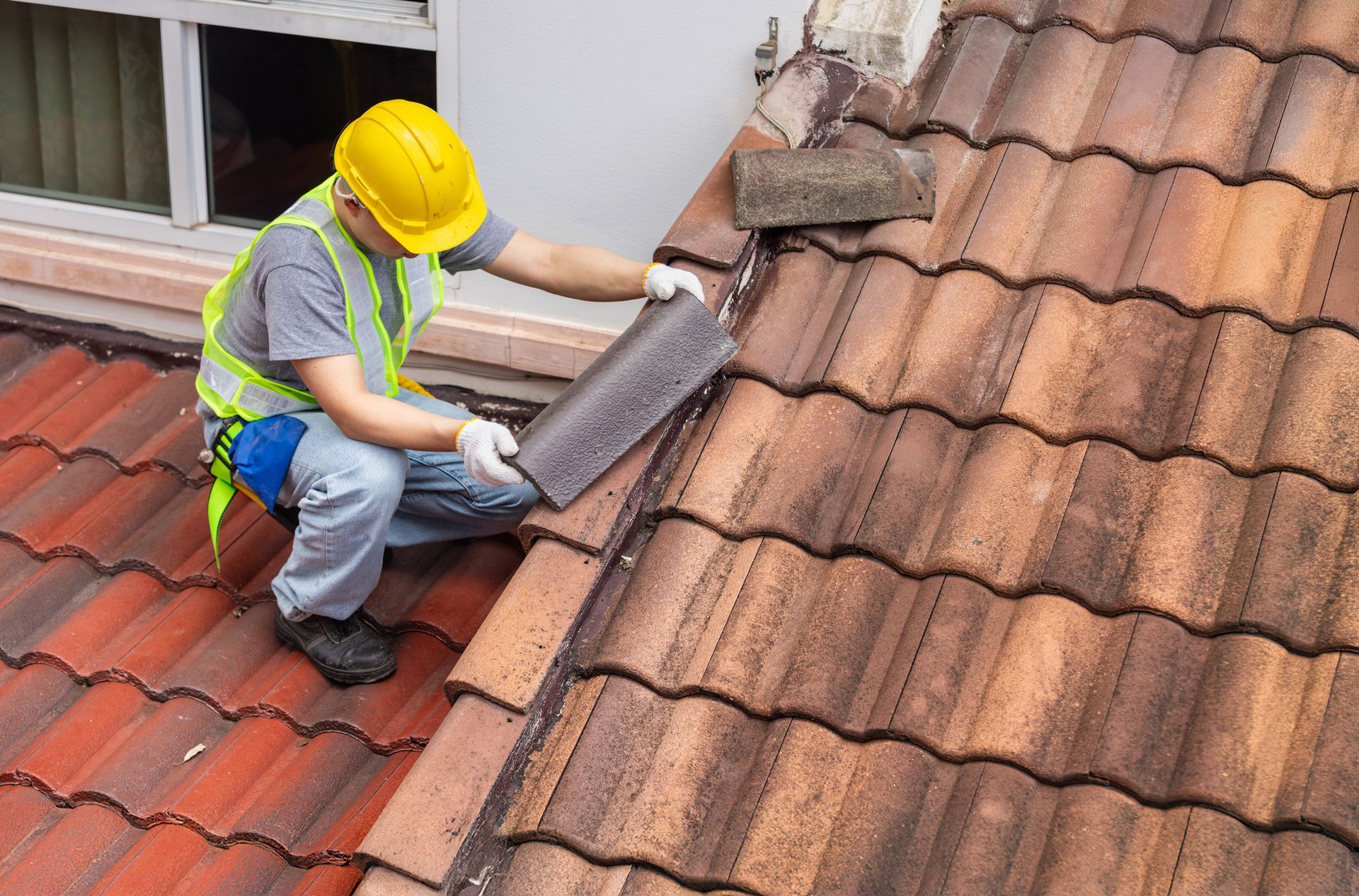 Roofer in hard hat and safety vest, installing a tile on a brown tiled roof.