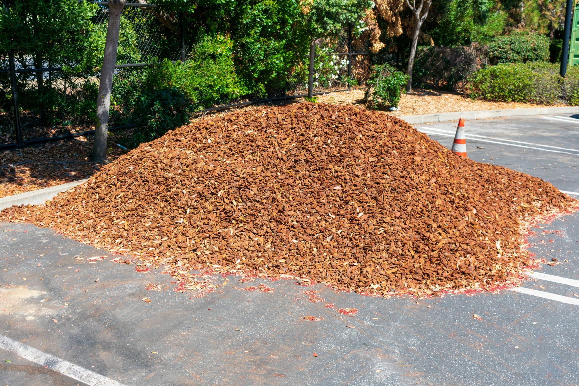 Large pile of brown mulch on an asphalt road beside trees and a traffic cone
