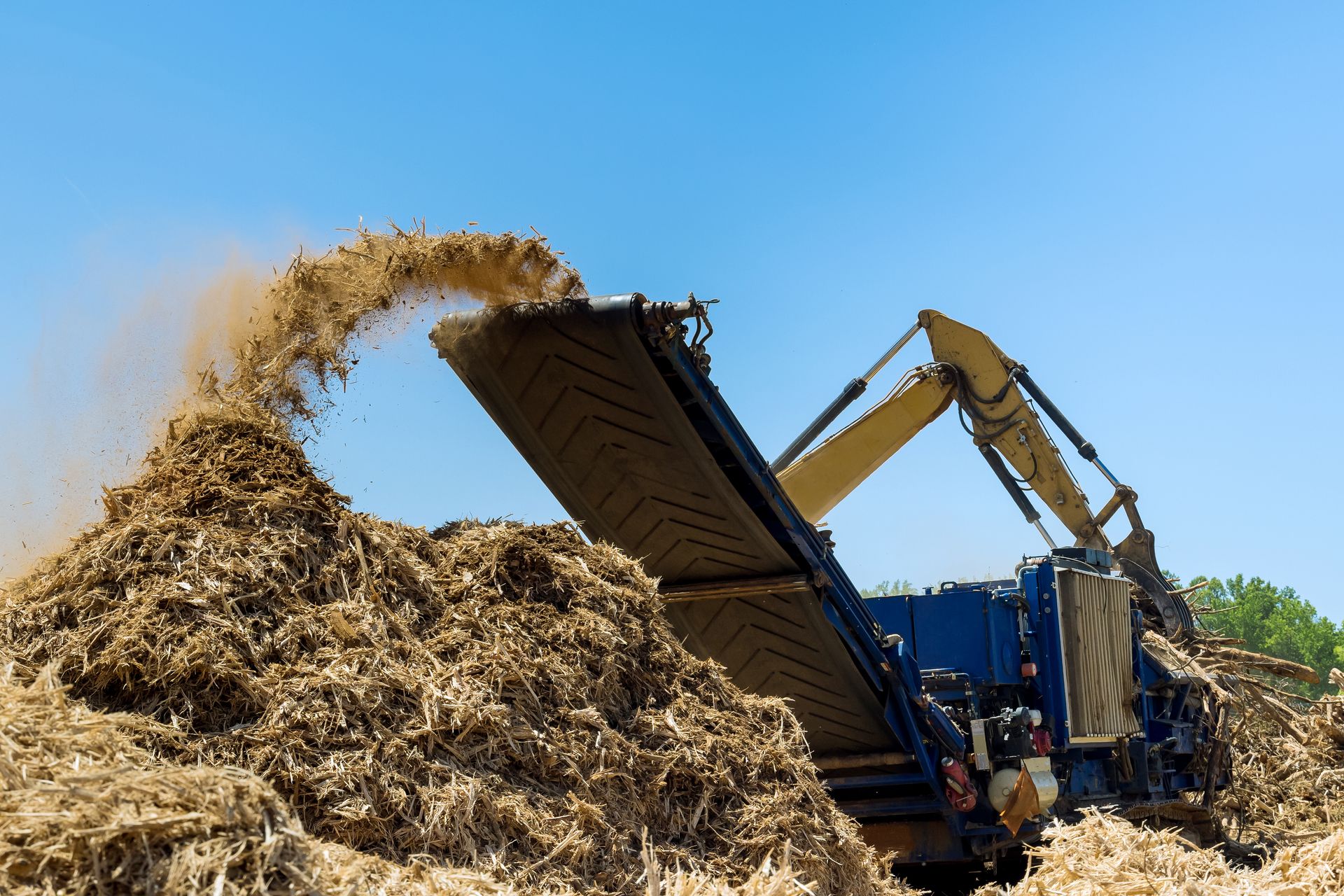 Excavator dumping wood chips into a large pile under a blue sky