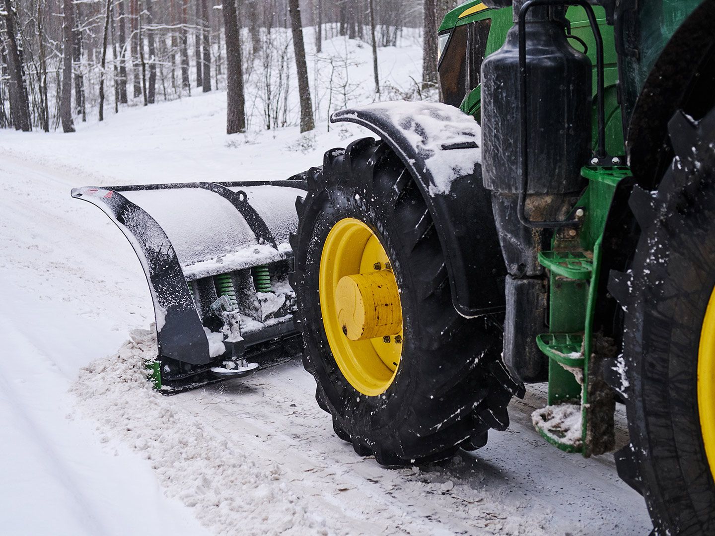 John Deere tractor plowing snow on a road with a black snowplow and yellow wheels.