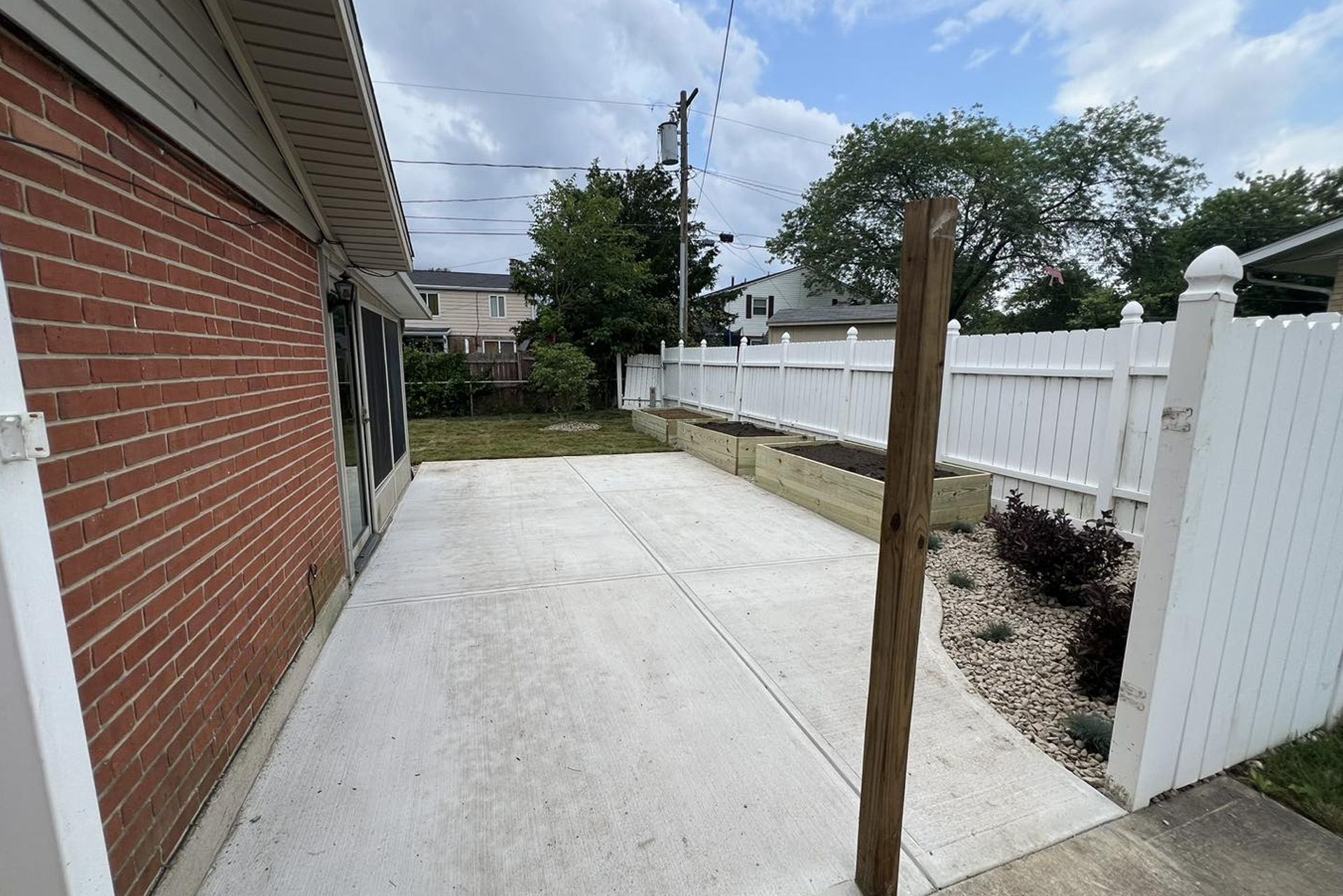 Concrete patio beside a red brick house, leading to a white fenced backyard with raised garden beds and plants.