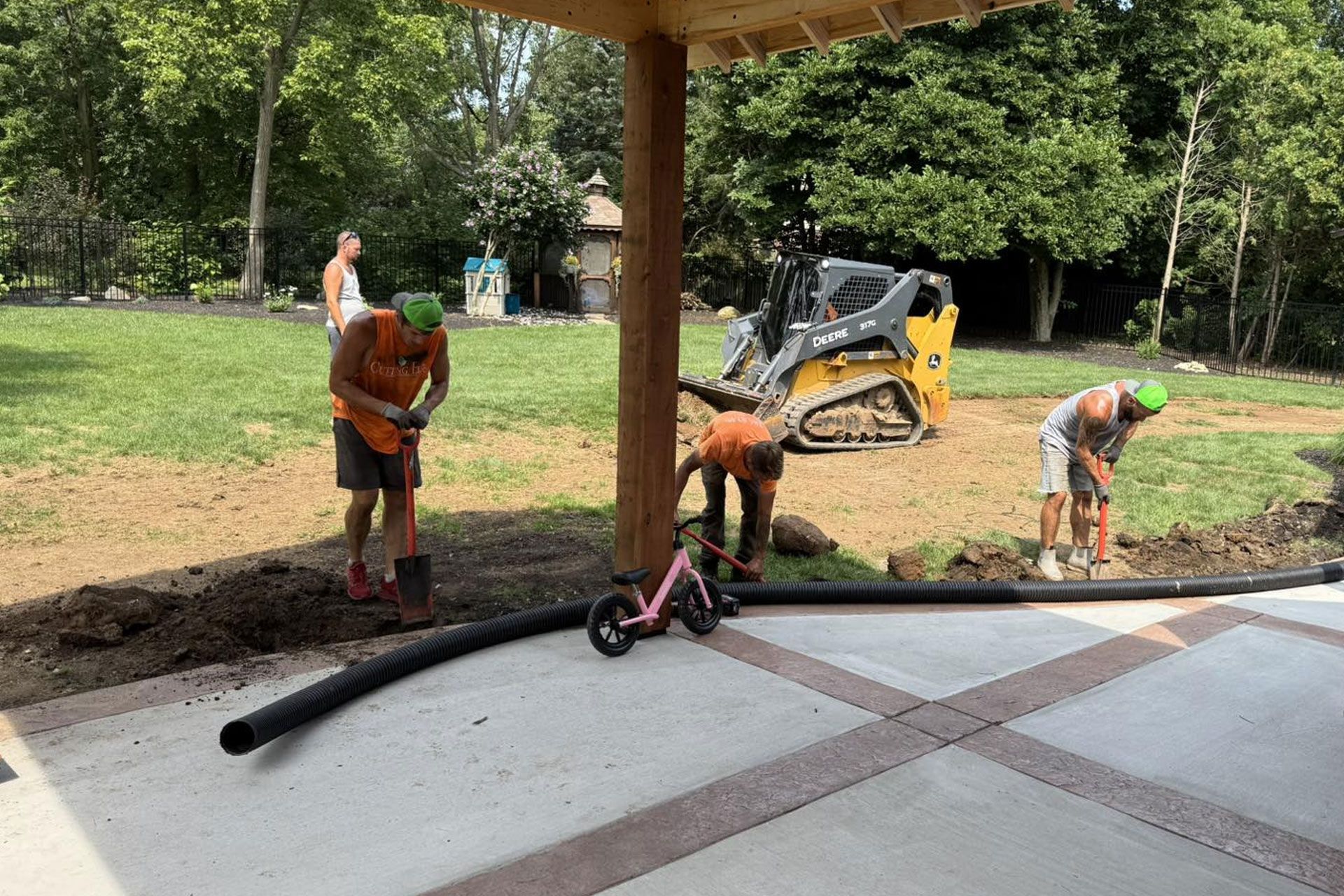 Construction workers installing drainage pipes near a patio and lawn, with a skid steer in the background.