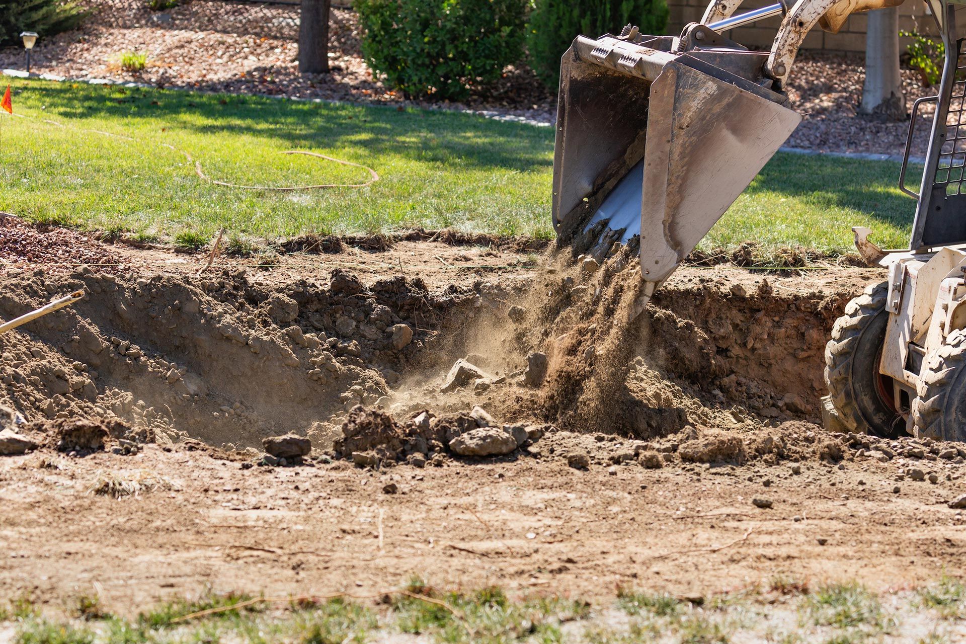 Skid steer bucket dumping dirt into a trench on a sunny lawn.