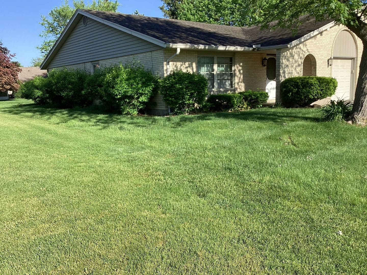 A green lawn in front of a tan-colored house with bushes and trees.