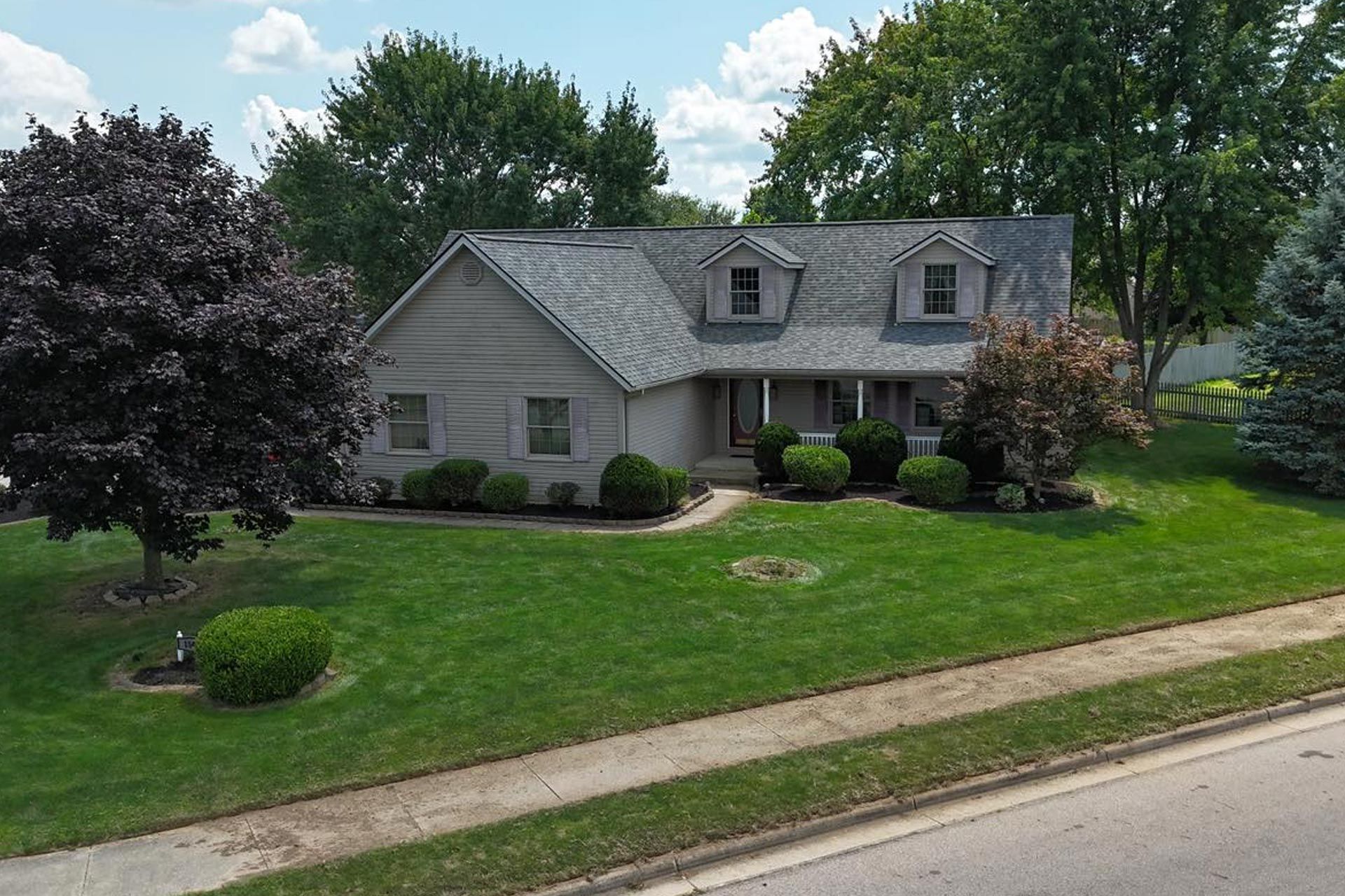 House with gray roof and siding, dormers, porch, surrounded by green lawn and trees.