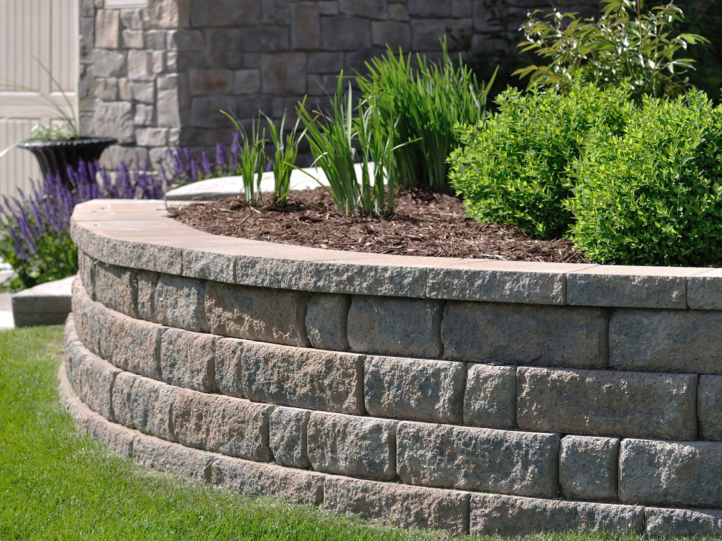 Curved retaining wall made of gray stone blocks with green plants and grass in a garden bed.