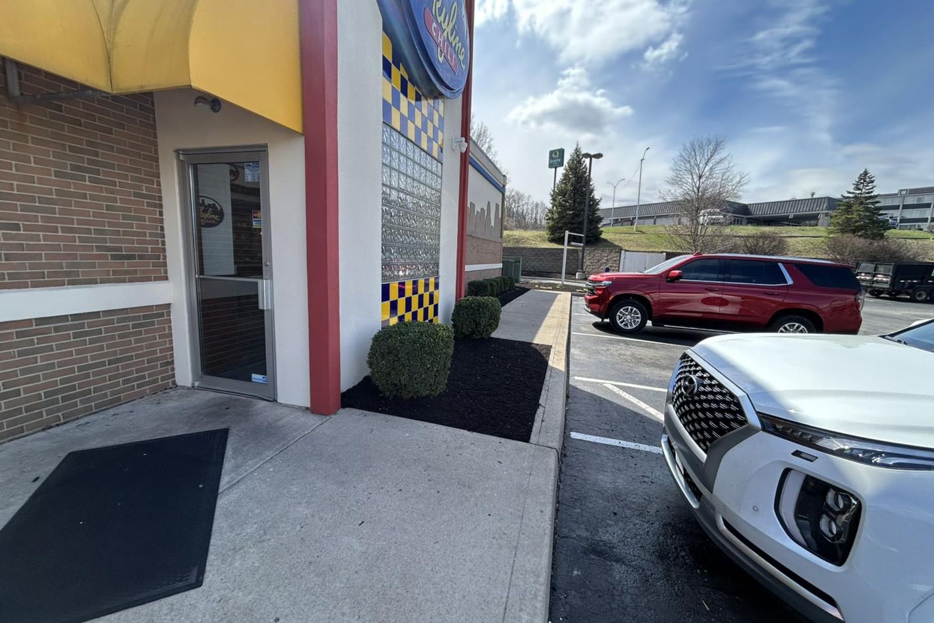 Exterior of a building with a yellow awning, red trim, and glass blocks. A red SUV and white car are parked nearby.