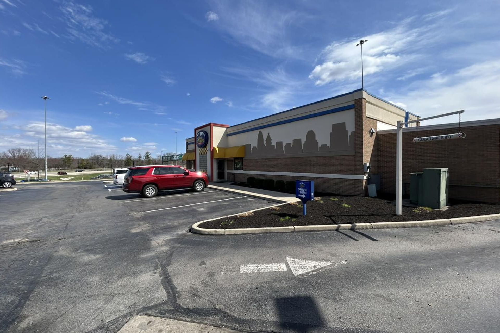 Red SUV parked in front of a fast-food restaurant with a skyline mural under a blue sky.