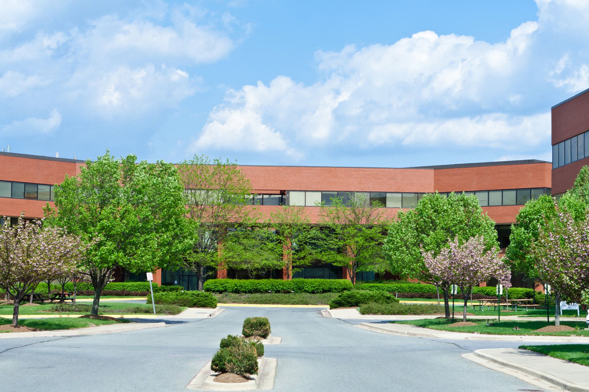 Brick office building with a curved facade, surrounded by trees and a paved driveway under a blue sky.