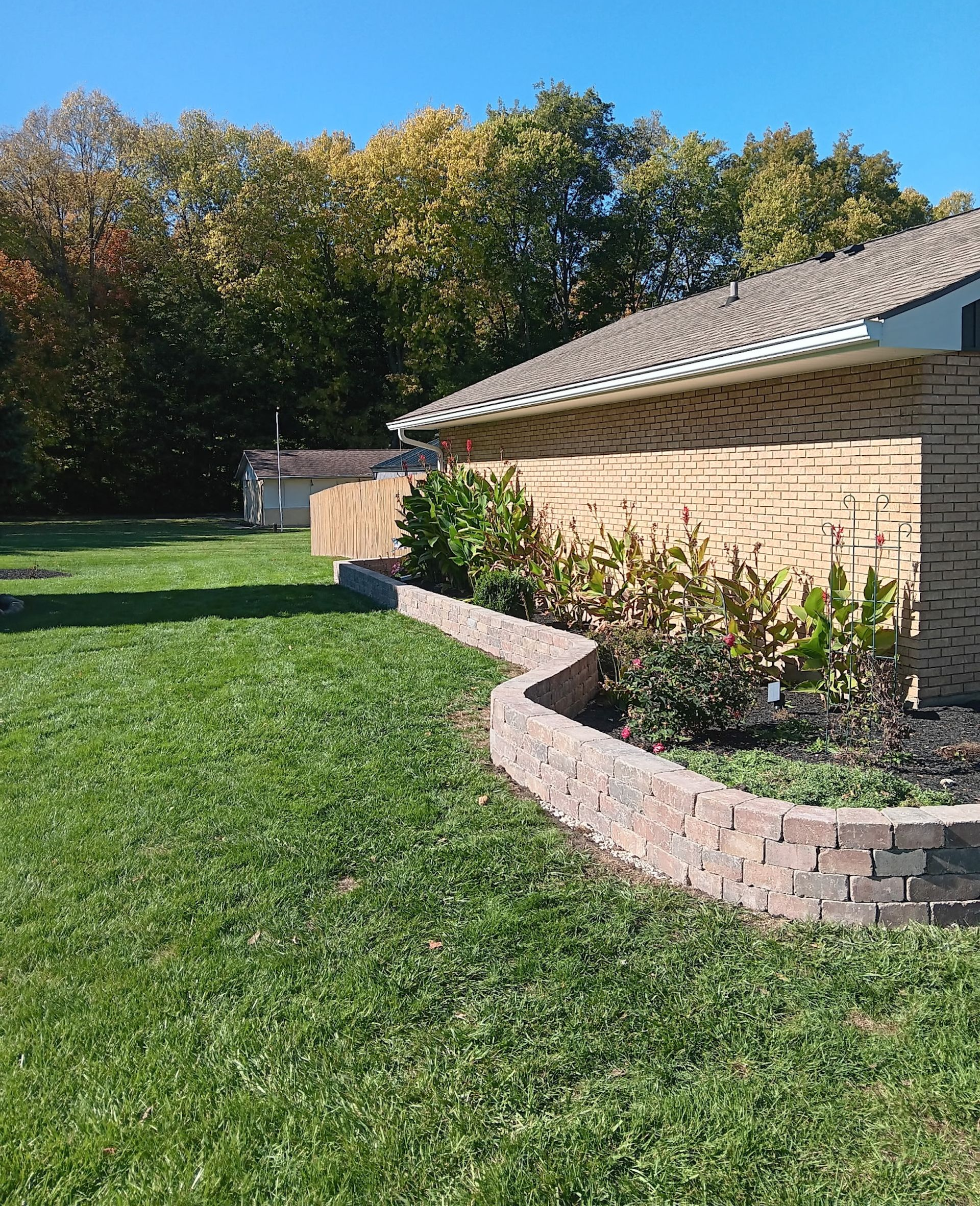 Brick retaining wall with plants, green lawn, and a brick building under a blue sky.