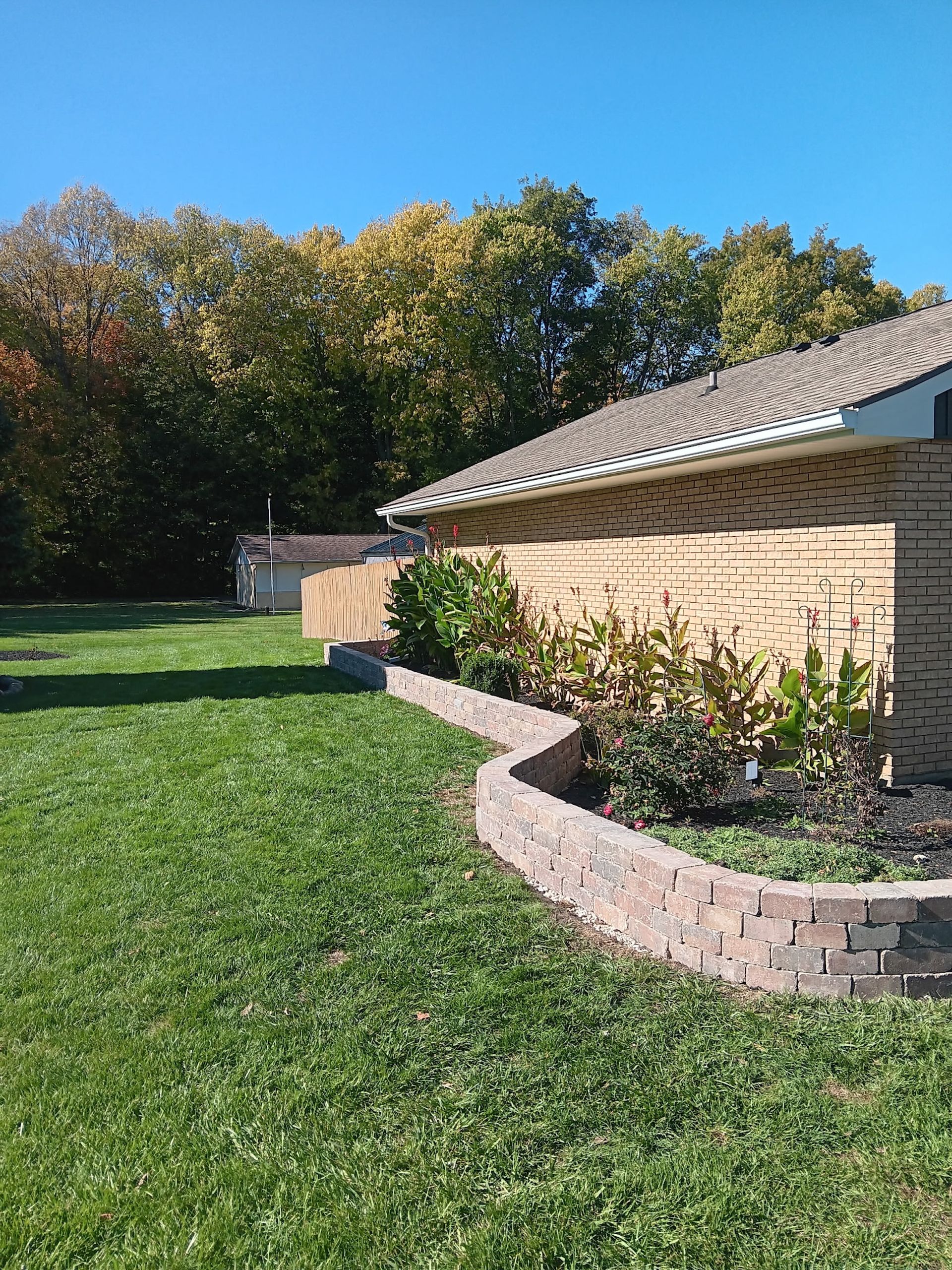 Backyard view of brick retaining wall with plants, building with tan brick, trees, and grass under a blue sky.