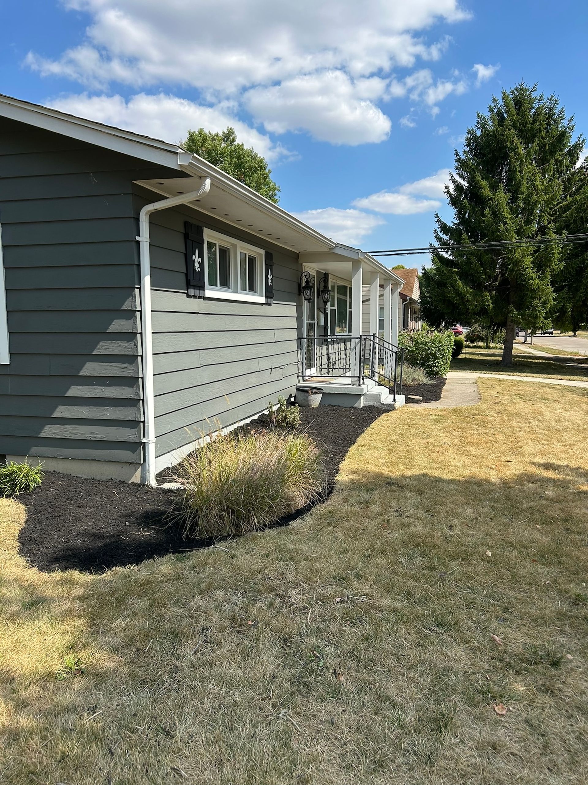 Gray house with black mulch, dry grass, and a sunny sky.