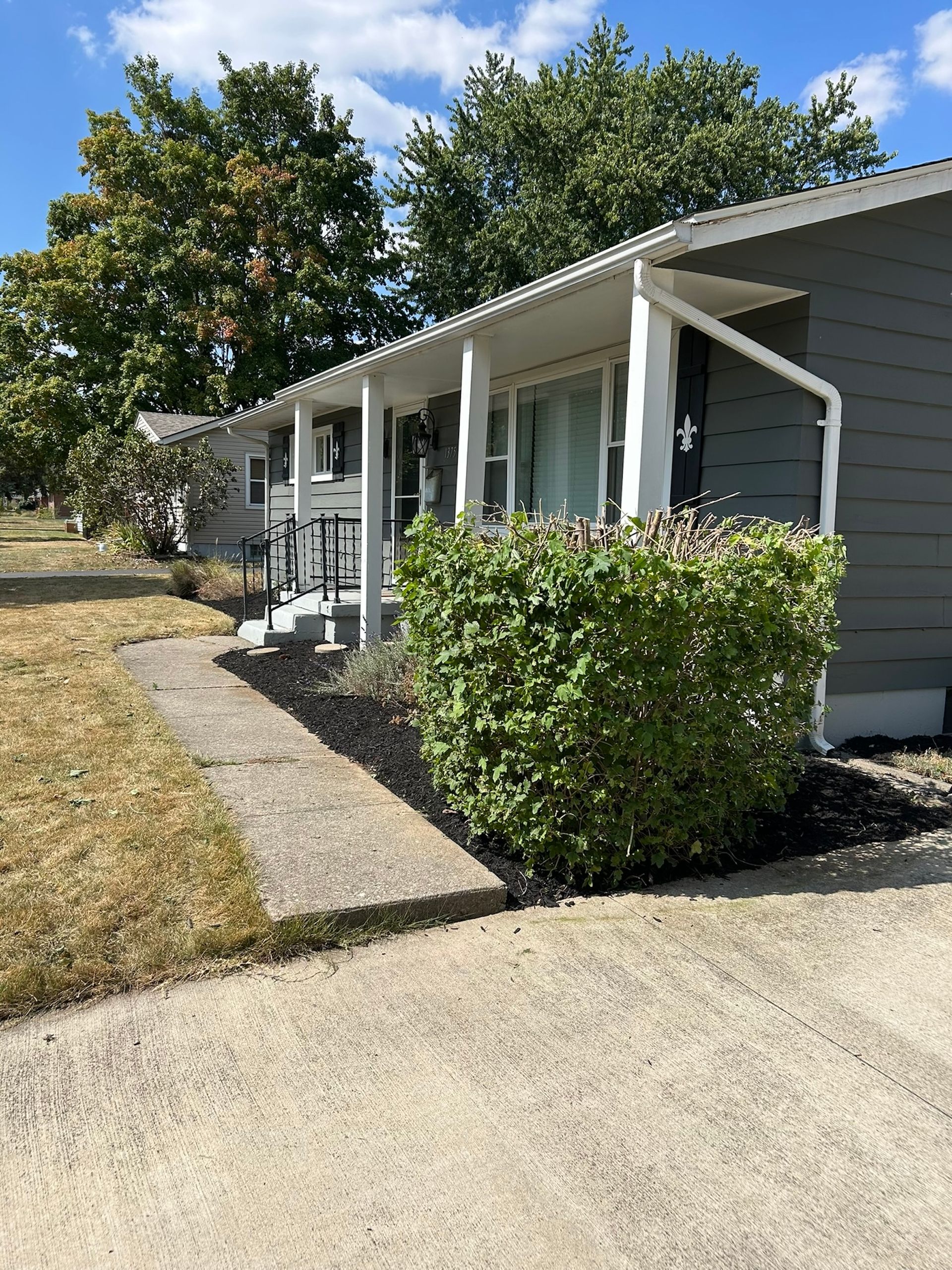 Gray building with white trim, porch, and a green hedge, set in a grassy yard.