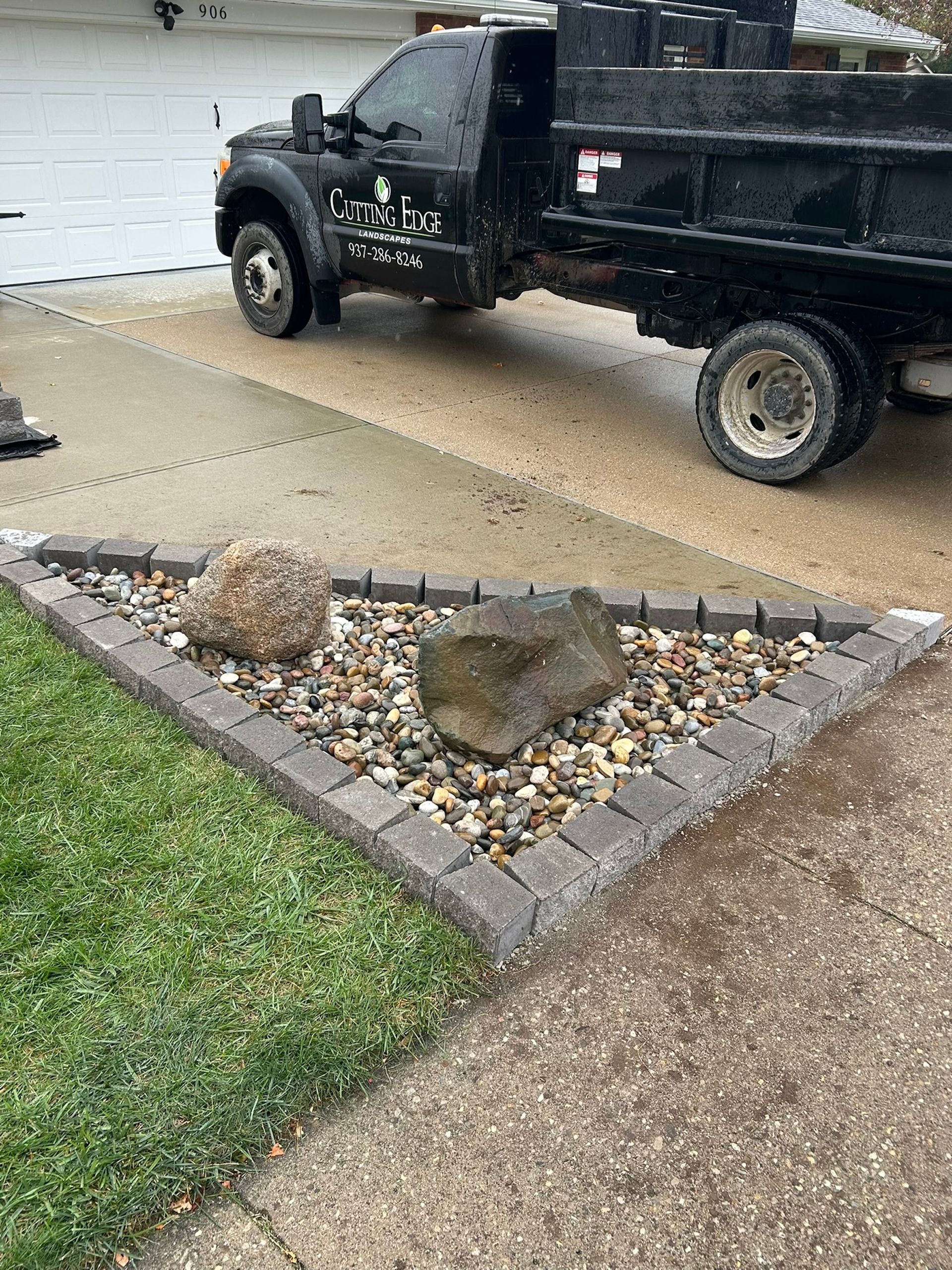 Triangular rock garden with a black dump truck in the background, set on a driveway next to a lawn.