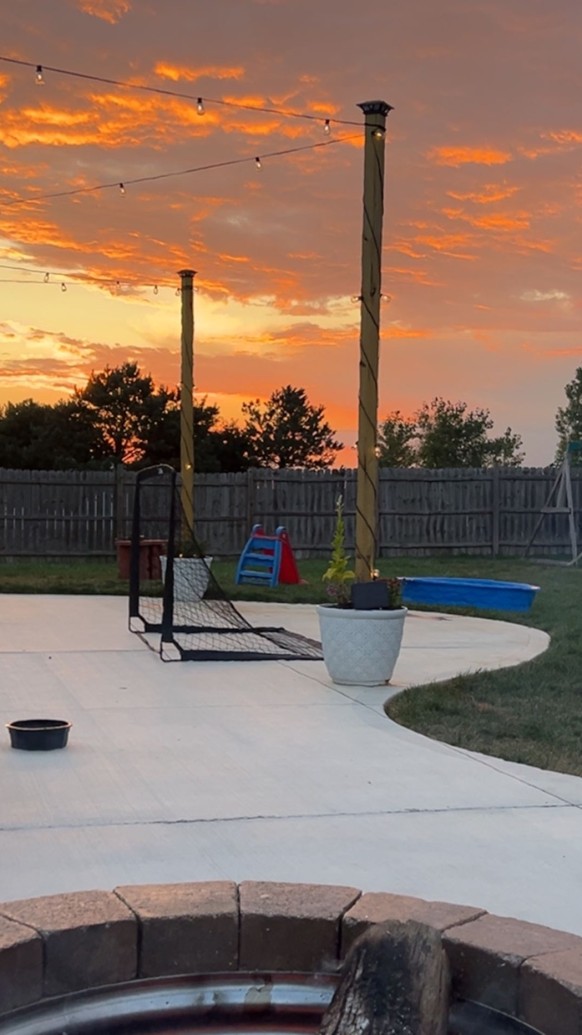 Backyard at sunset, concrete patio, two wooden poles, string lights, blue kiddie pool, orange sky.