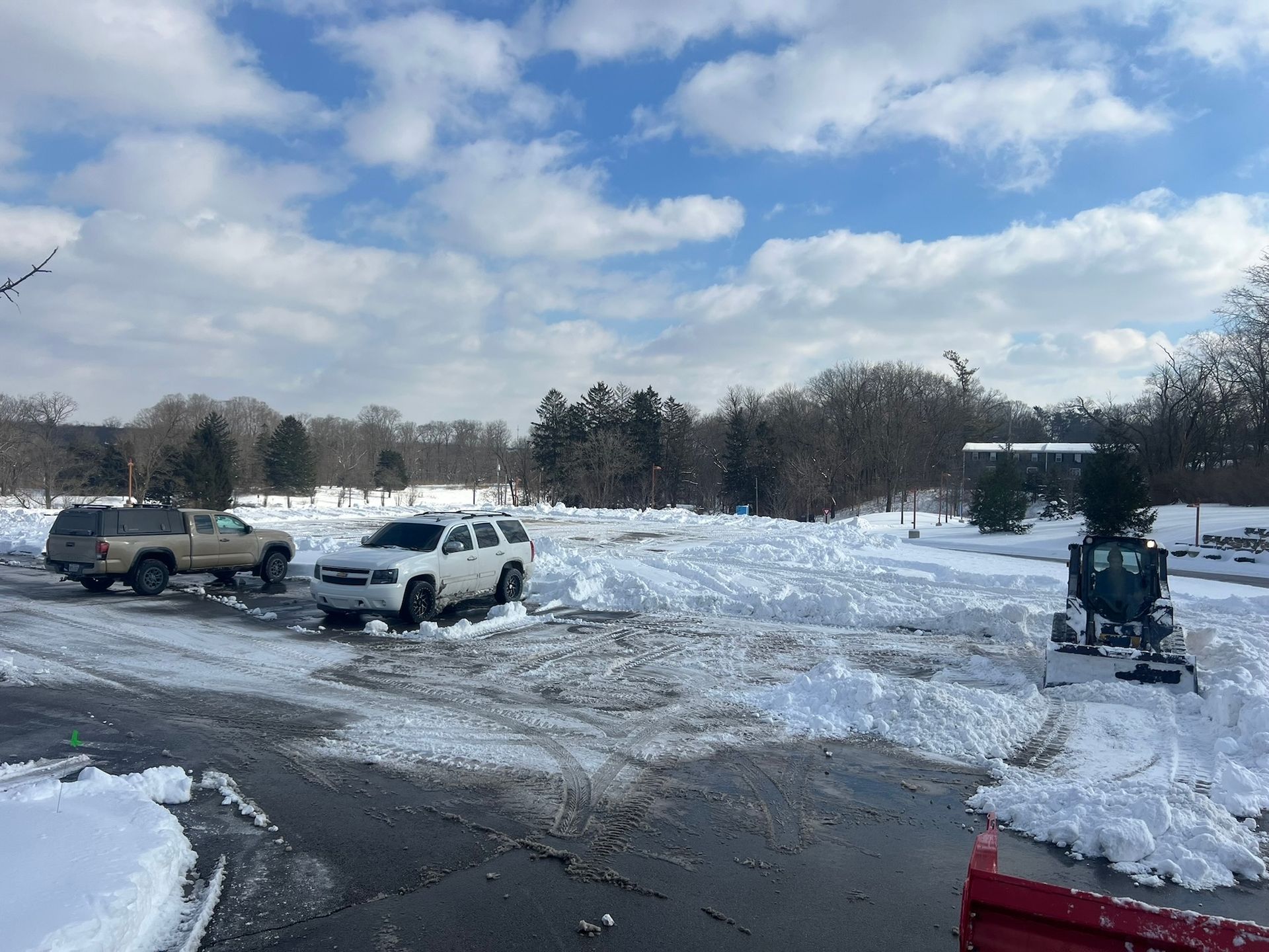 Snow-covered parking lot with trucks and a snowplow removing snow. Sunny, daytime. Trees in the background.