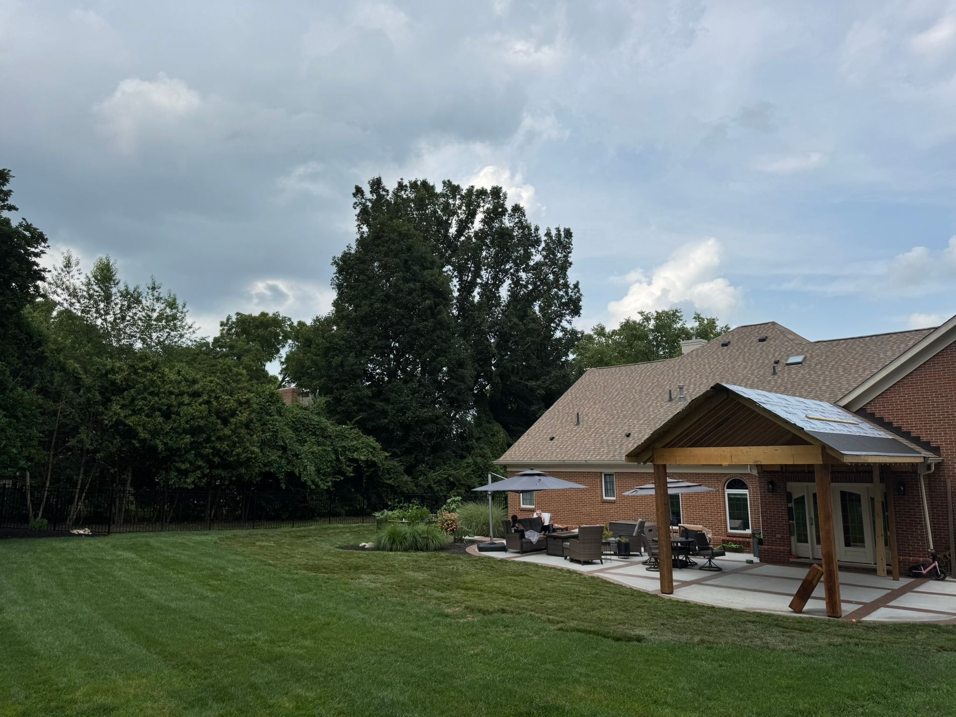 Backyard with lawn, patio, brick house, and trees under a cloudy sky.