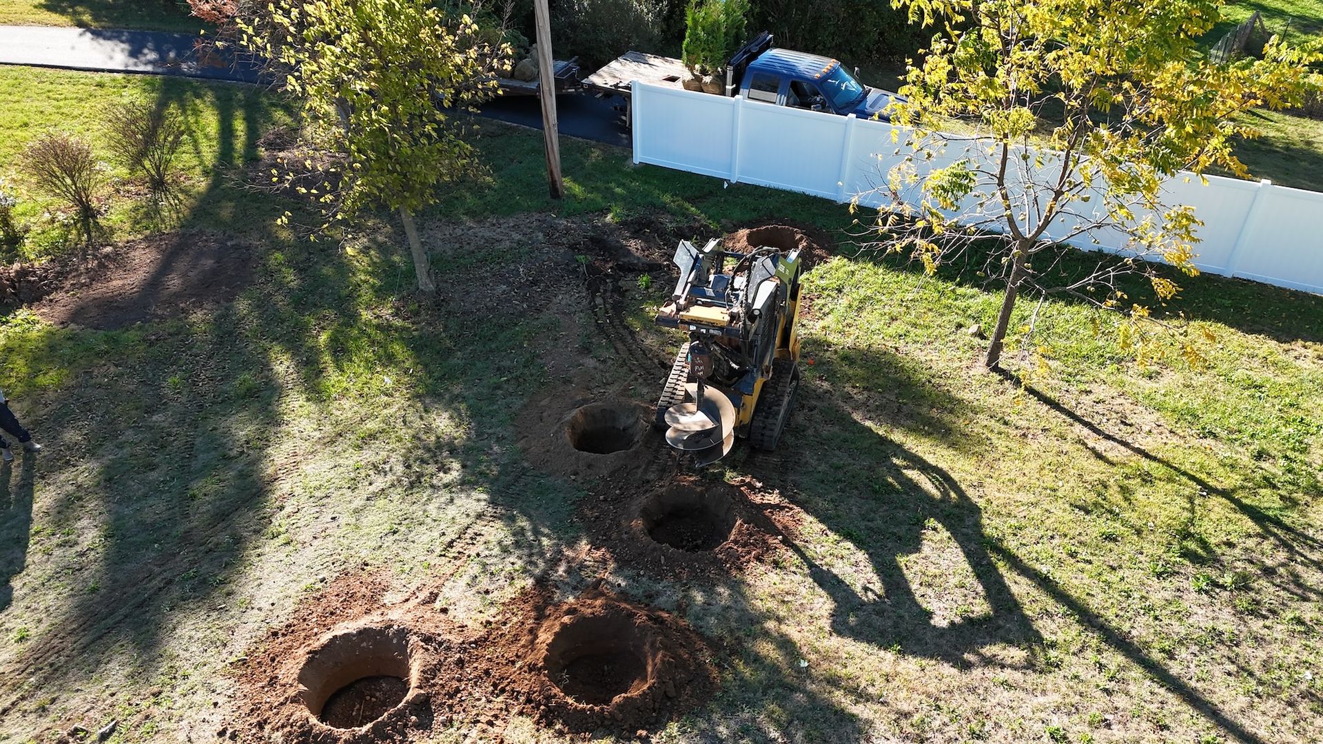 Skid steer digging holes in a grassy yard, near a white fence, possibly for planting trees.