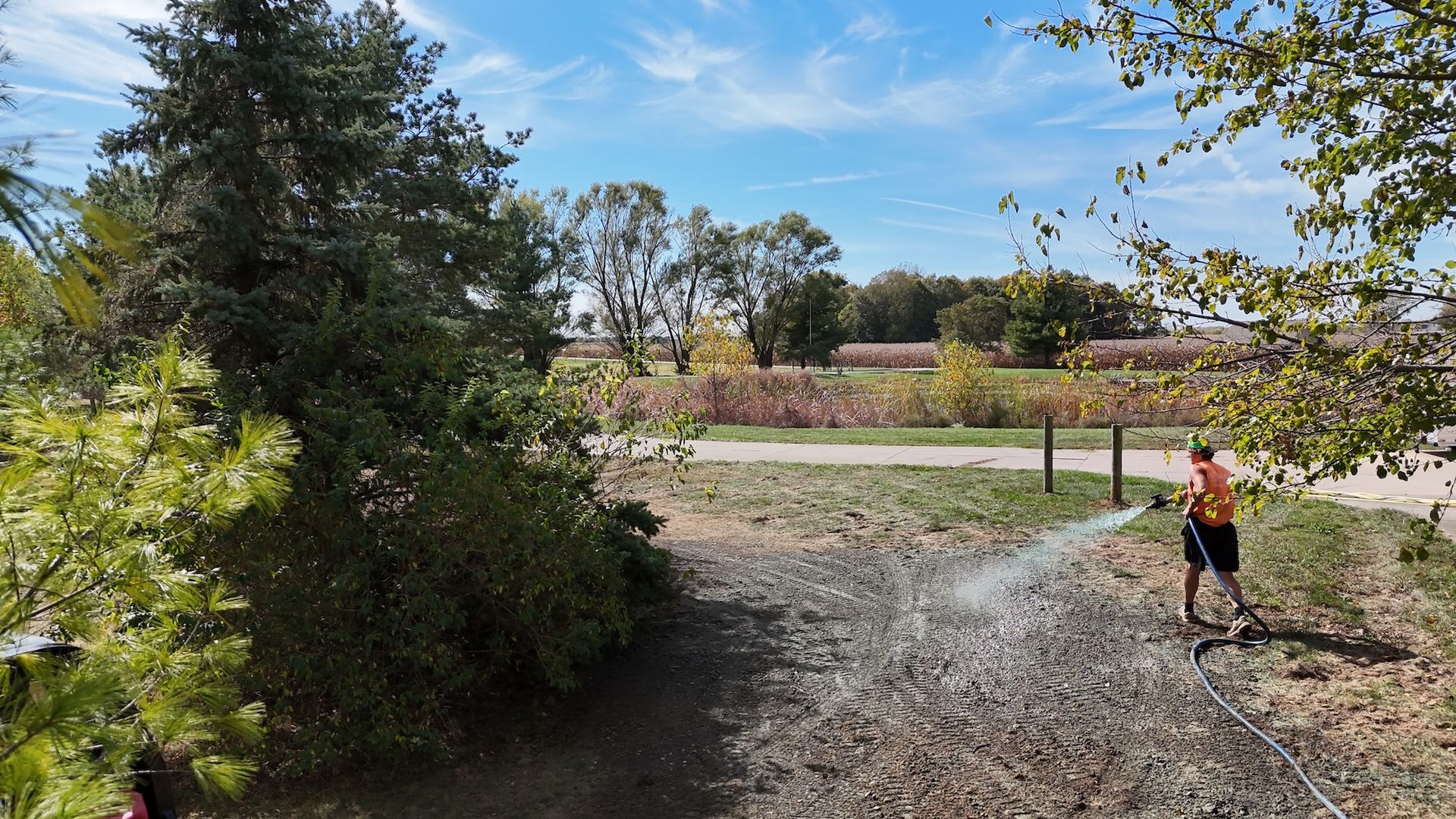 Person watering a dirt path in a sunny outdoor setting with trees, a fence, and fields in the background.