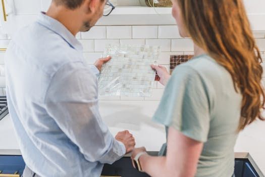 Couple examining a floor plan in a kitchen, man in blue shirt, woman in green, white tile backsplash.