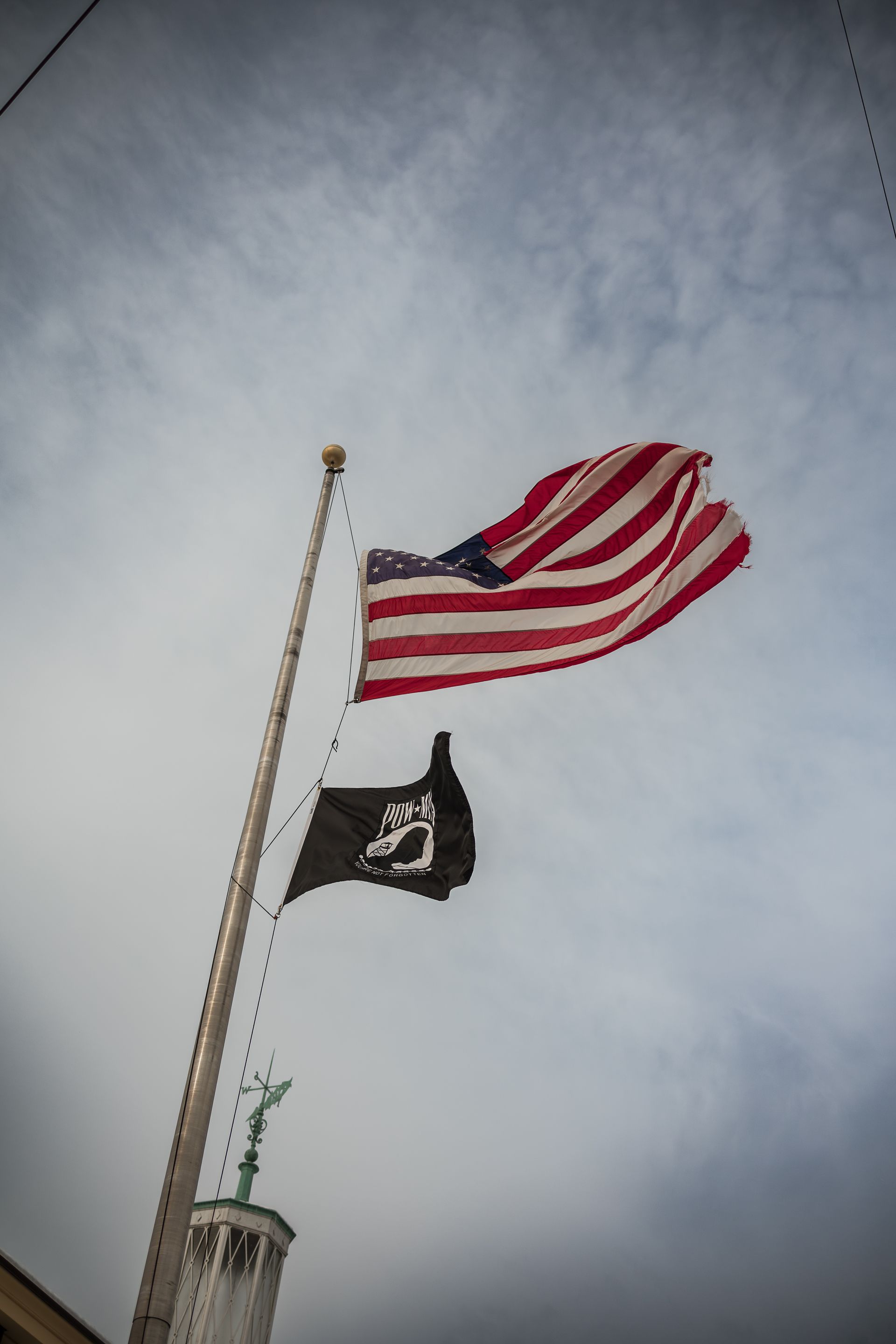 American flag draped over a dark coffin with white flowers. Person in black suit in background.