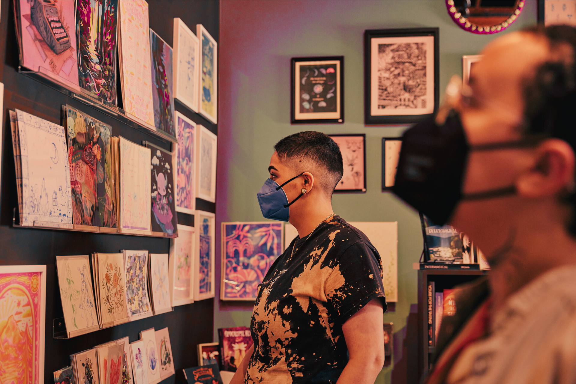 Close-up side view of two masked non-binary friends scanning a wall filled with colorful framed illustrations. A Latinx person with a crew cut fade and ear gauges is centered in frame. In the right foreground, a Black person with glasses and noise-canceling headphones is visible, though blurred because of depth of field.
Link Here: https://affecttheverb.com/gallery/disabledandhere/gallerywall/