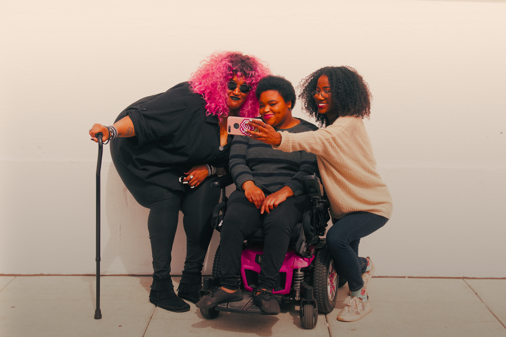 A zoomed out full body shot showing three Black and disabled friends (a non-binary person with a cane and tangle stim toy, a non-binary person sitting in a power wheelchair, and an invisibly disabled femme) smiling and taking a cell phone selfie together. All are outdoors in front of a white wall.
Image Credit: https://affecttheverb.com/collection/