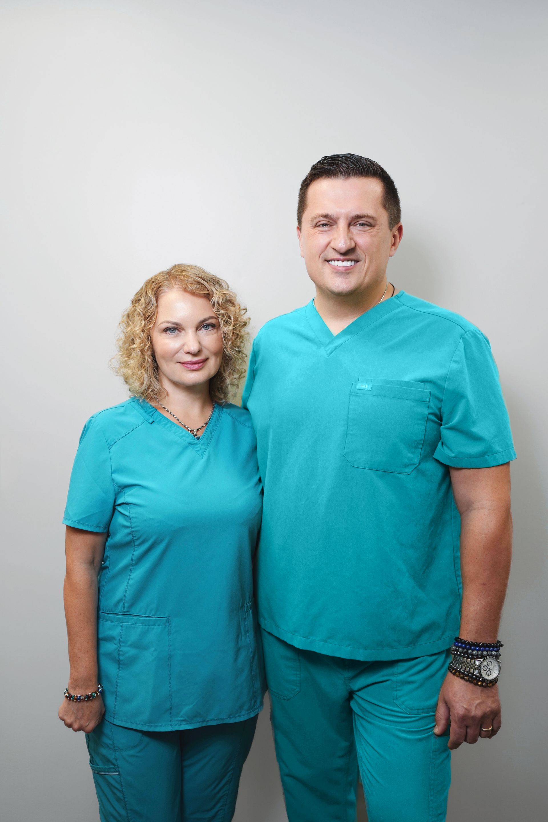 Two medical professionals in turquoise scrubs smile at the camera.