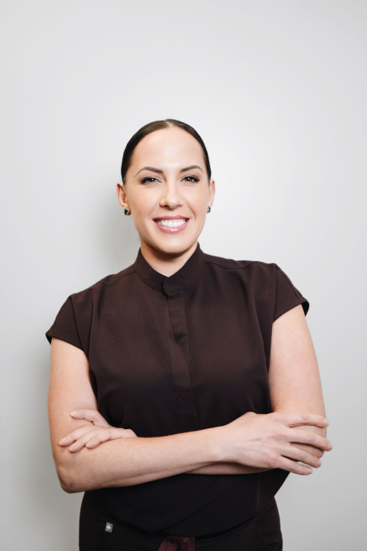 Woman with arms crossed, smiling, wearing a brown top, in front of a white wall.
