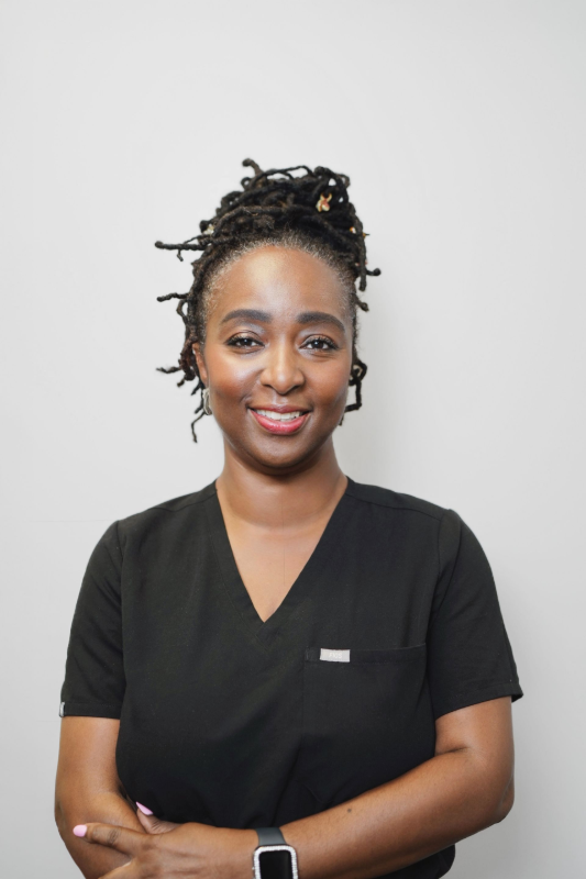 Woman with locs wearing black scrubs, smiling, indoors.