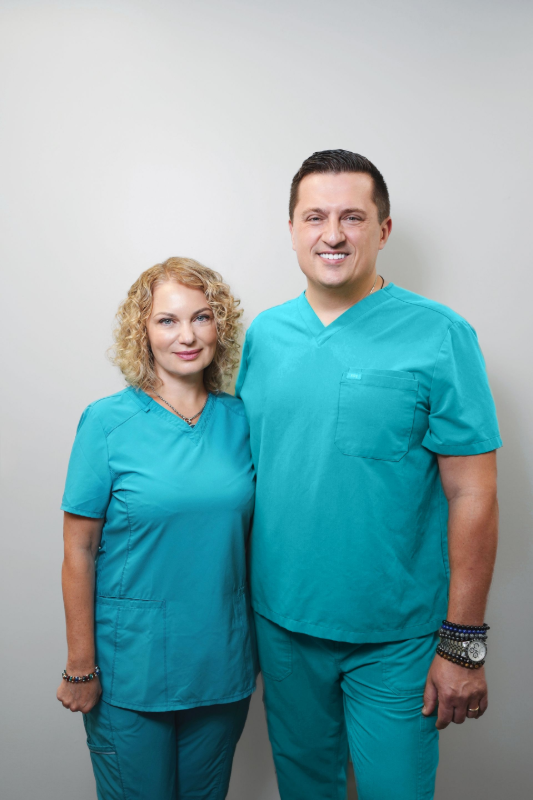 Two people in teal scrubs: a woman with blonde curly hair and a man. They smile, posing against a white background.