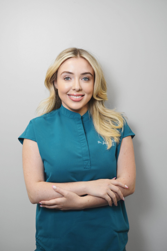 Woman with blonde hair, wearing teal scrubs, smiling with arms crossed, in front of a gray wall.