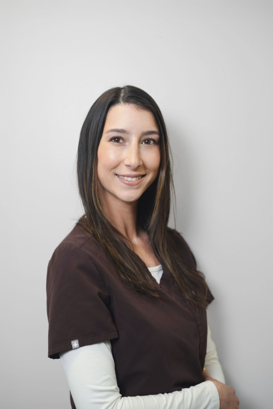 Woman in brown scrubs smiles at the camera, standing against a white wall.