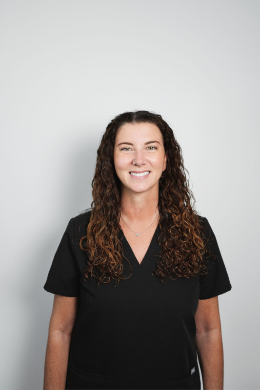 Woman with brown curly hair smiles, wearing a black v-neck top, against a white background.